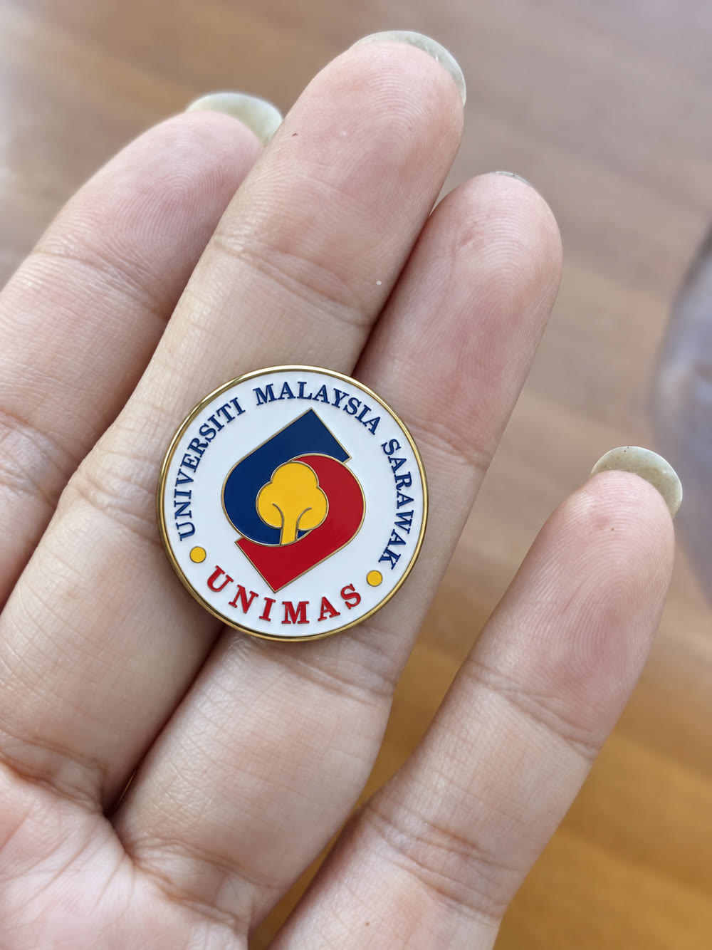Round gold-rim enamel lapel pin with the UNIMAS crest, held between fingers